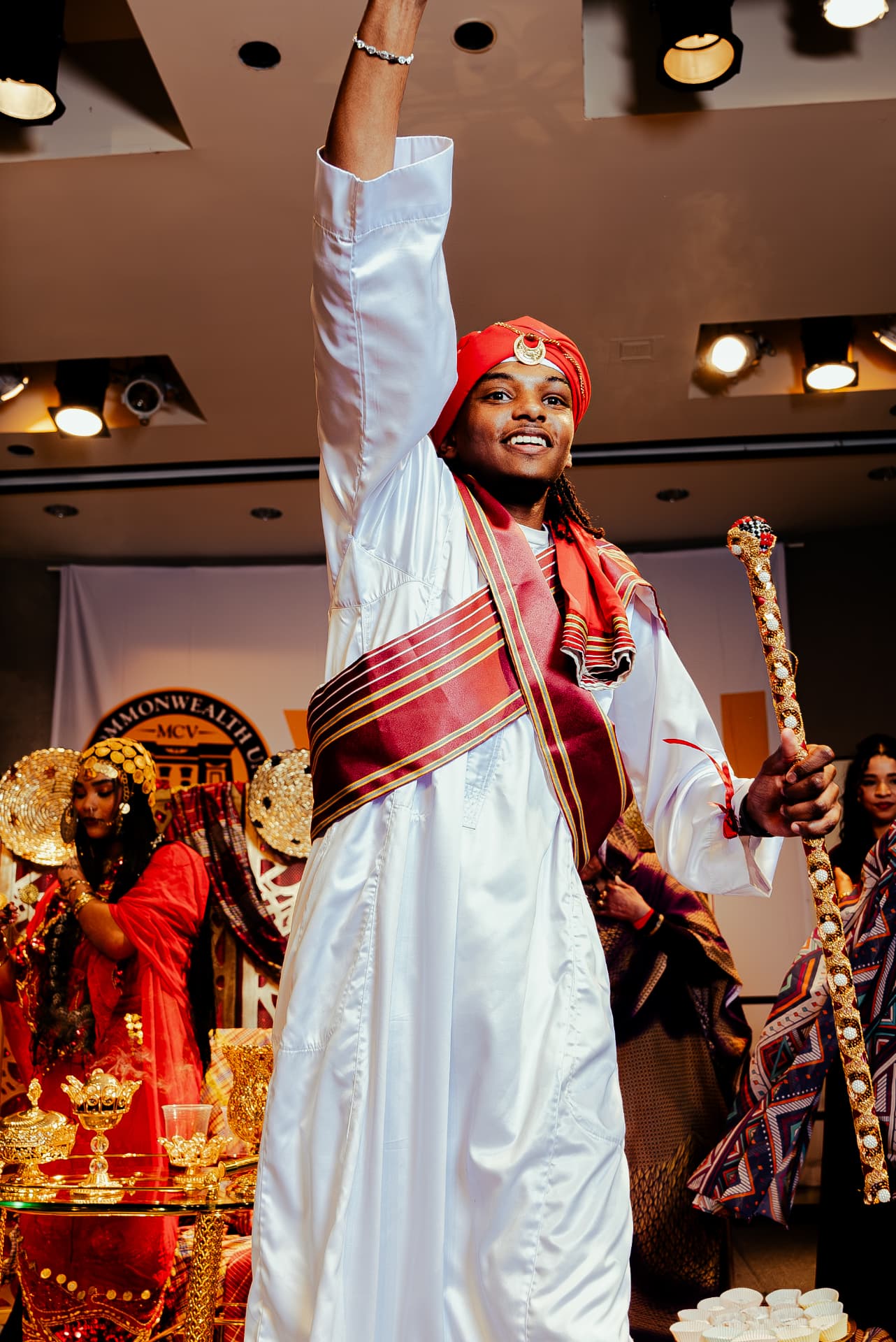 Student in white jalabiya and red turban raising his hand at Taste of Sudan